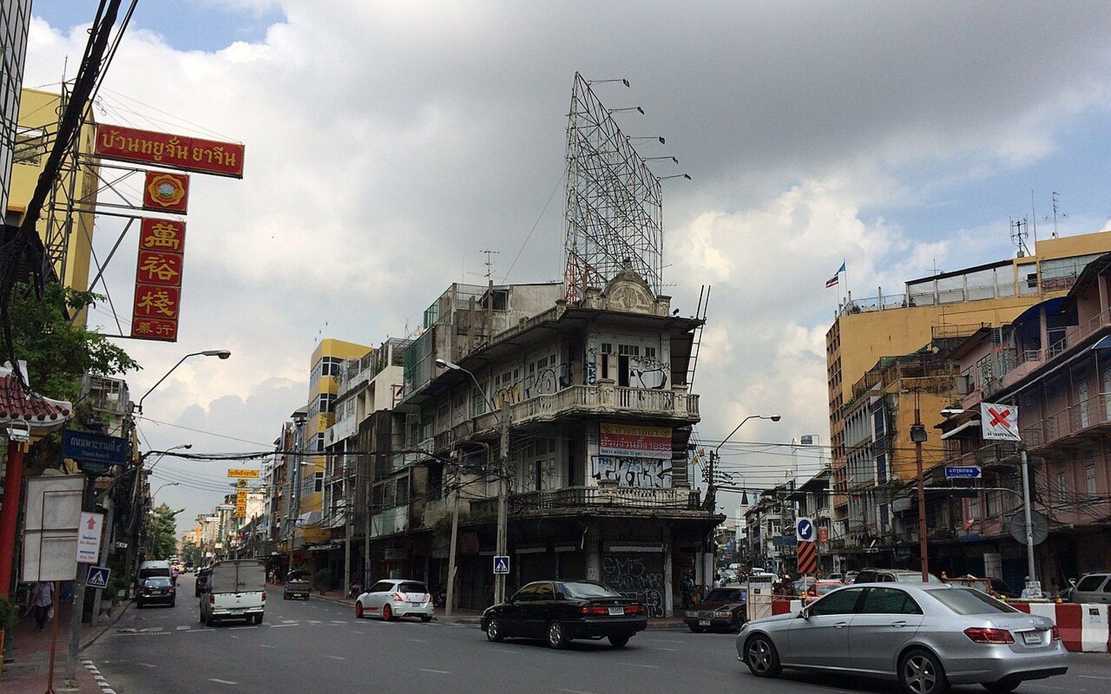 Traffic passes a corner building with a rooftop billboard frame under a cloudy sky