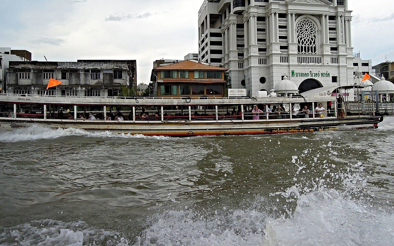 A crowded long-tail passenger boat speeds along a river past waterfront buildings in Bangkok