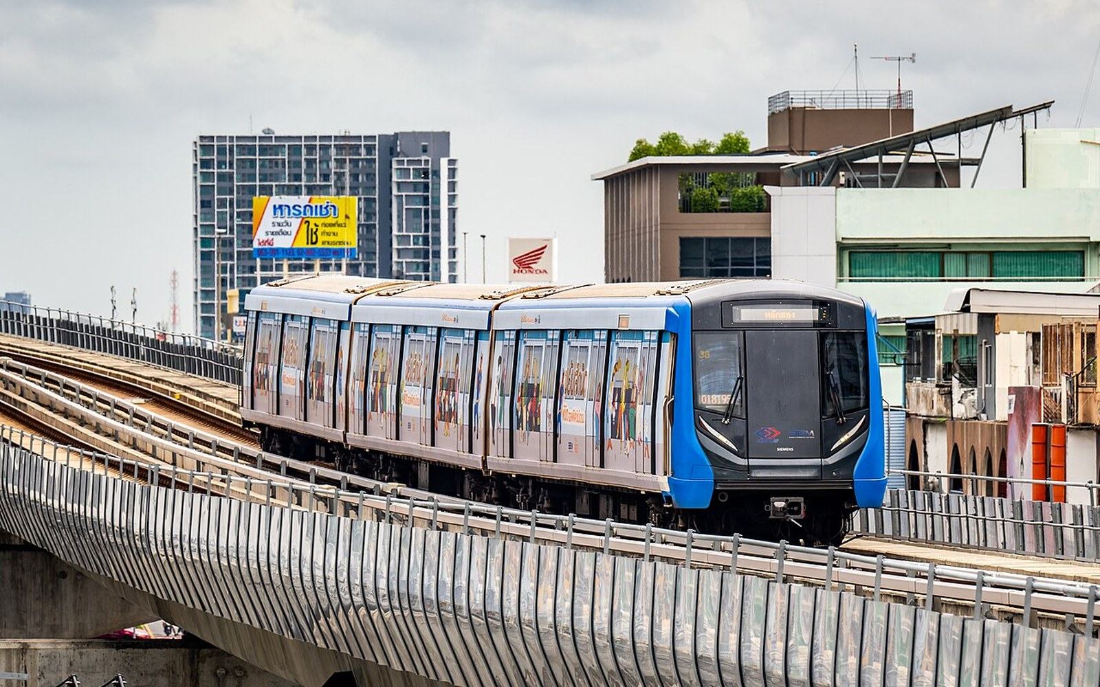 An elevated train travels along an urban rail line above city buildings and signage