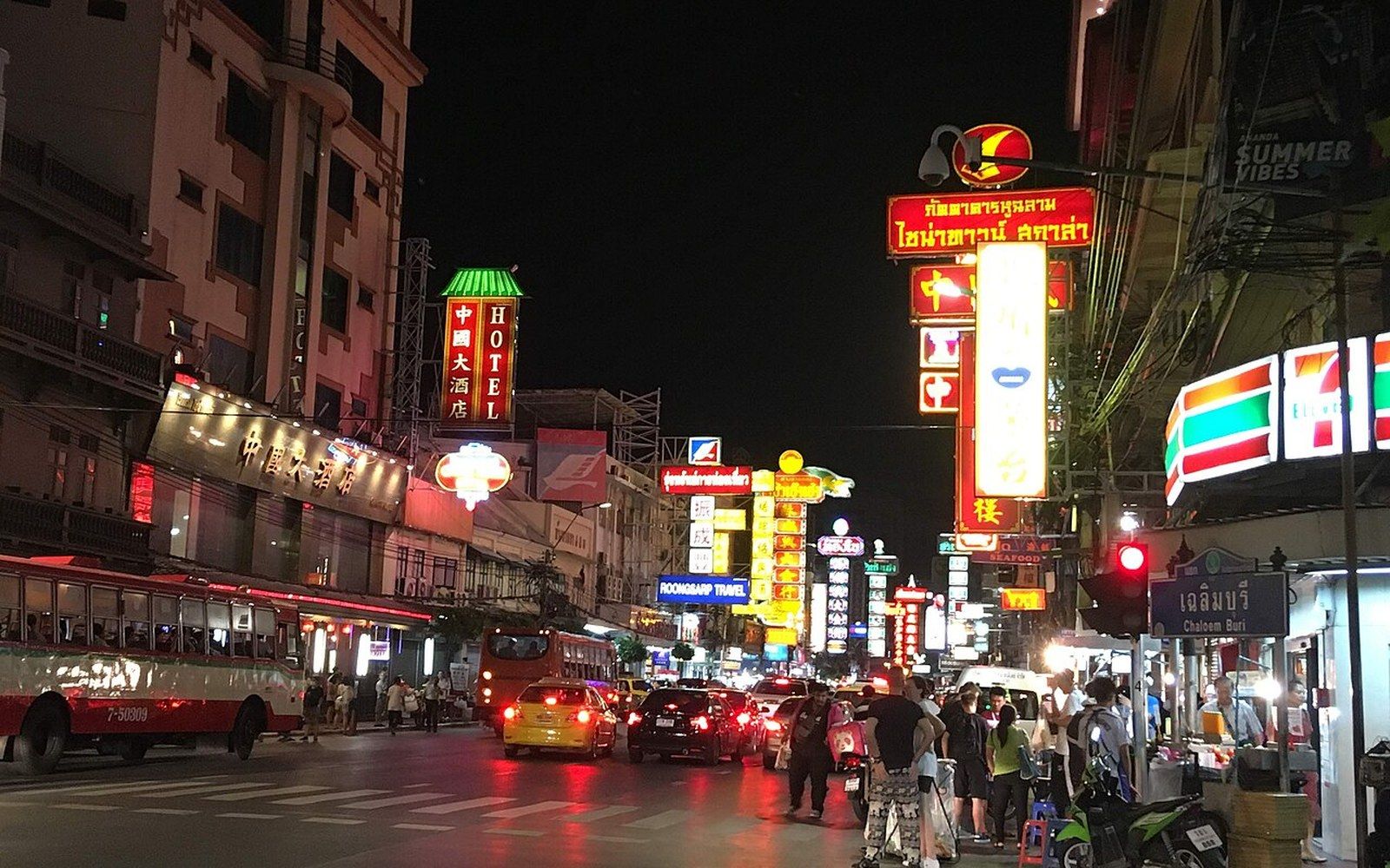 Neon signs light up a busy street in Bangkok's Chinatown at night with traffic and pedestrians
